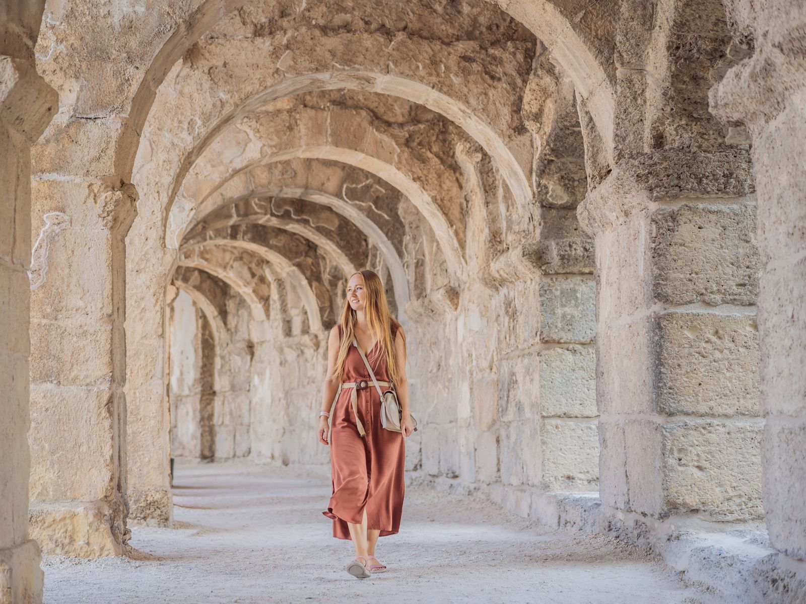 Девушка в терракотовом платье гуляет по древнему каменному коридору с арками в Аспендосе